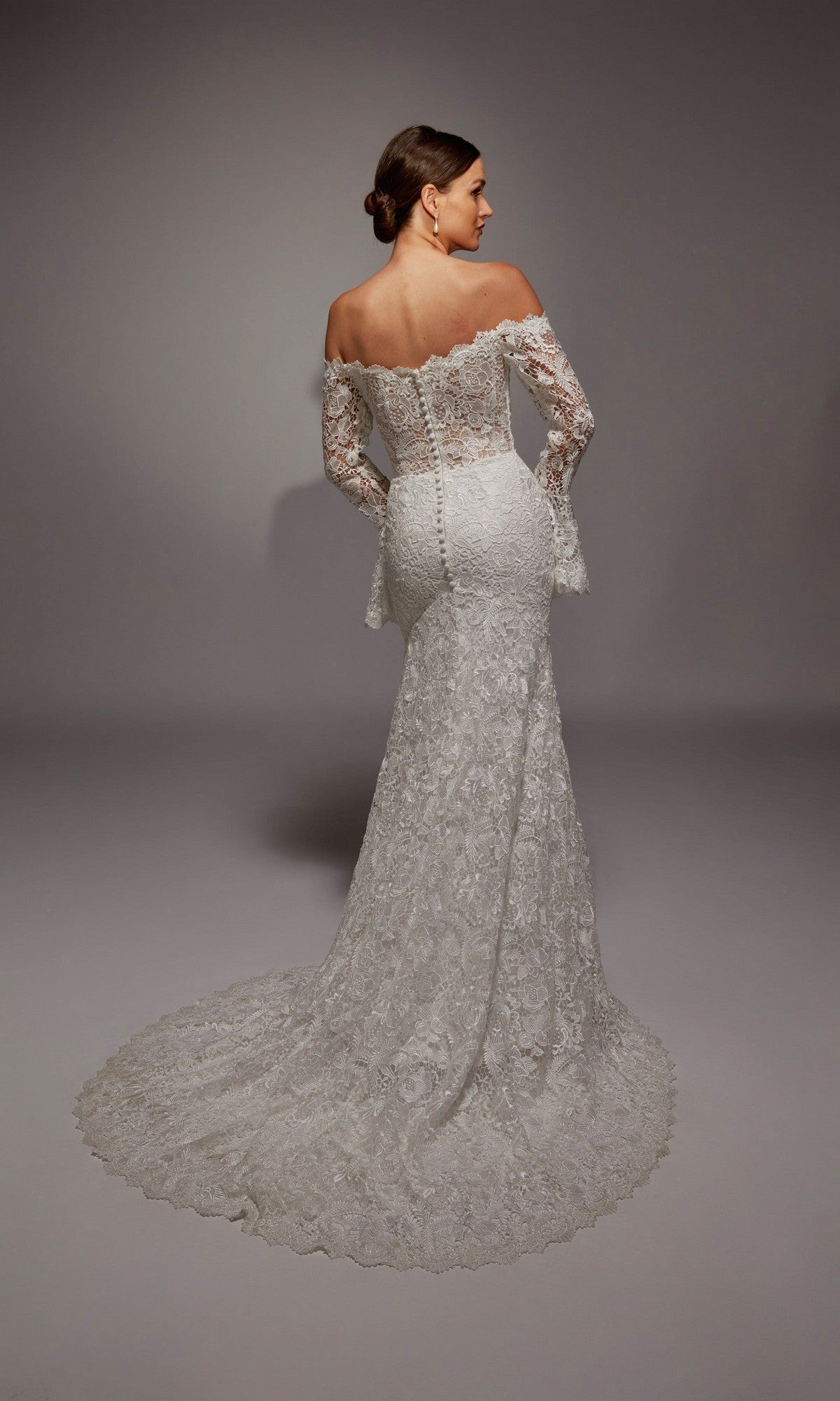 A woman stands against a plain background in the Alyce 7156: a long white, vintage-inspired bridal gown featuring an off-the-shoulder neckline, long lace sleeves, buttoned back, and a sweeping train.